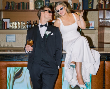 girl in white dress sitting on bar with man in tuxedo looking at her